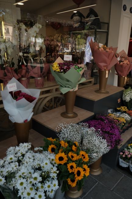 A floral display featuring three large bouquets of fresh flowers arranged in tall, cone-shaped brown paper wrapping, placed on wooden and elevated black platforms inside a flower shop. The bouquets in