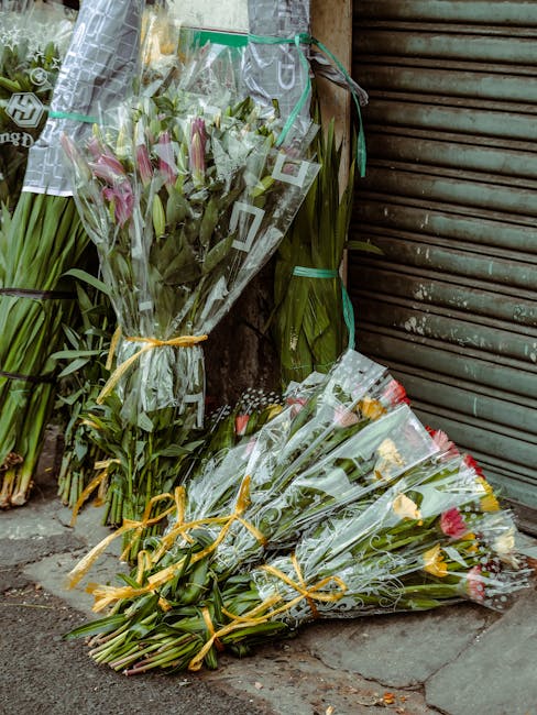 Several bouquets of fresh flowers, including pink lilies with delicate petals and green foliage, are wrapped in clear cellophane and tied with yellow ribbons. One bouquet is leaning against a weathere