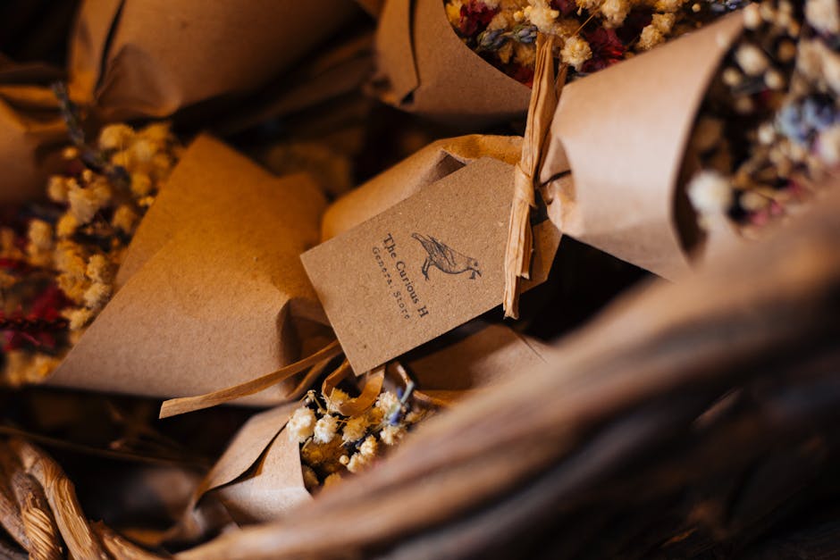 Close-up of a bouquet of dried flowers wrapped in brown kraft paper, with a small tag displaying a hummingbird illustration and the words 'The Curious Bird Est. 2016'. The arrangement features various