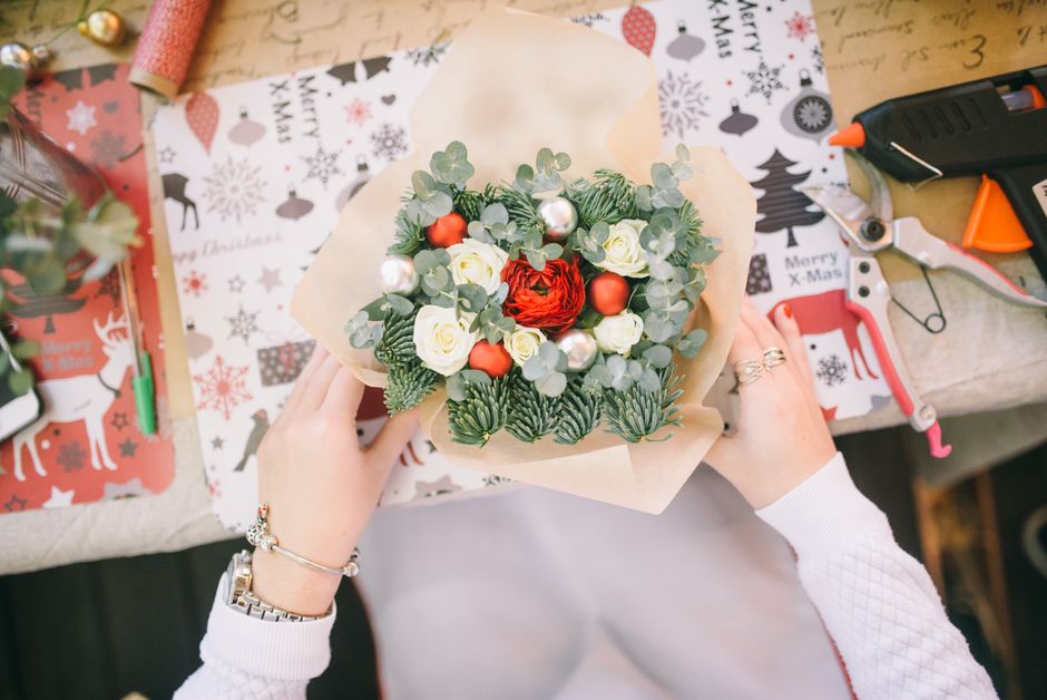A florist at a work table holding a festive floral gift arrangement featuring white roses, red ranunculus, eucalyptus leaves, and small orange berries, all wrapped in beige parchment paper. The arrang