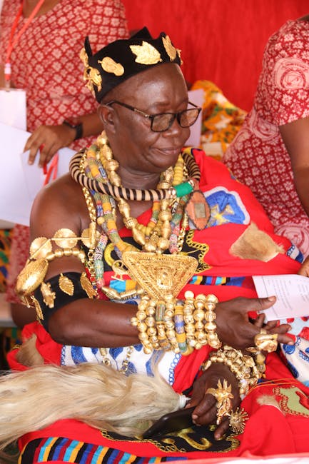 The image features a woman dressed in vibrant traditional African attire, adorned with numerous gold and beaded jewelry, including necklaces, bracelets, and a large gold pendant. She is wearing a deco